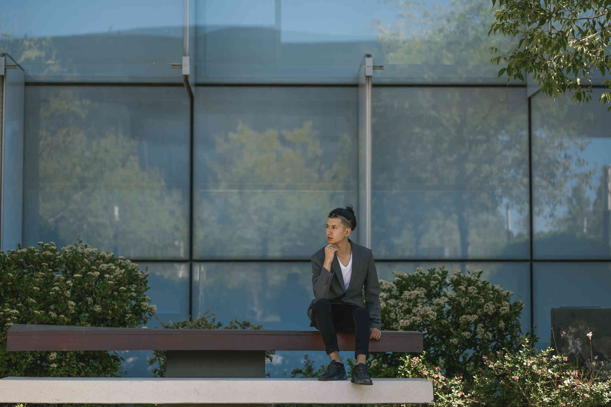 A man in a blazer sits outside on a picnic table near a glass building as he gazes off deep in thought.