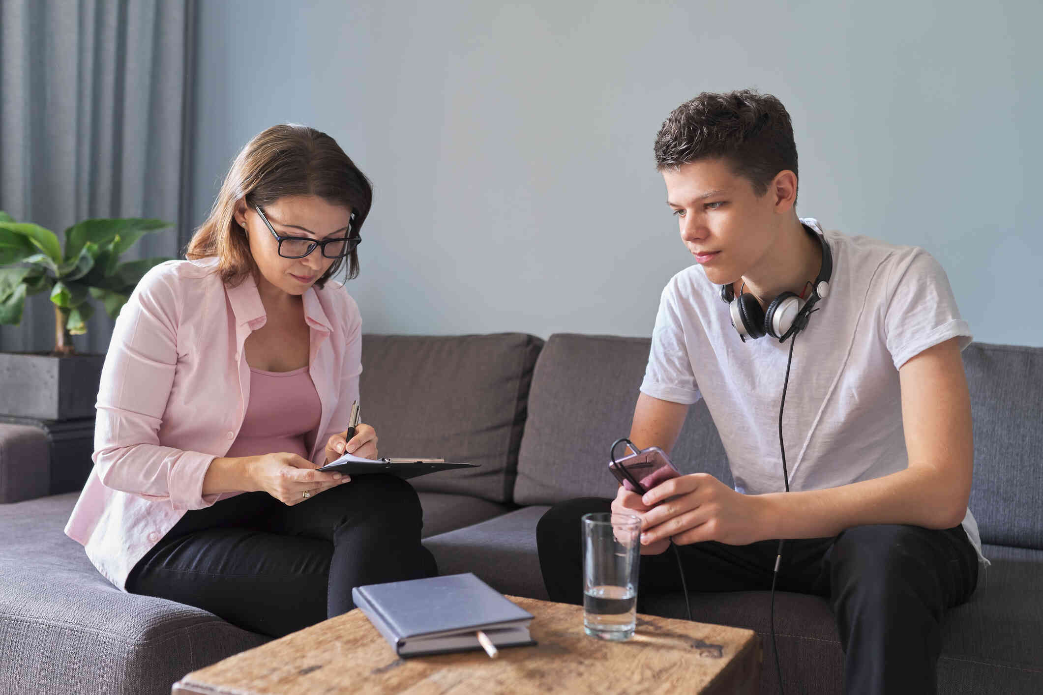 A young male teenager with headphones sits on a couch, engaging in a conversation with a woman taking notes, suggesting a counseling or mentoring session.