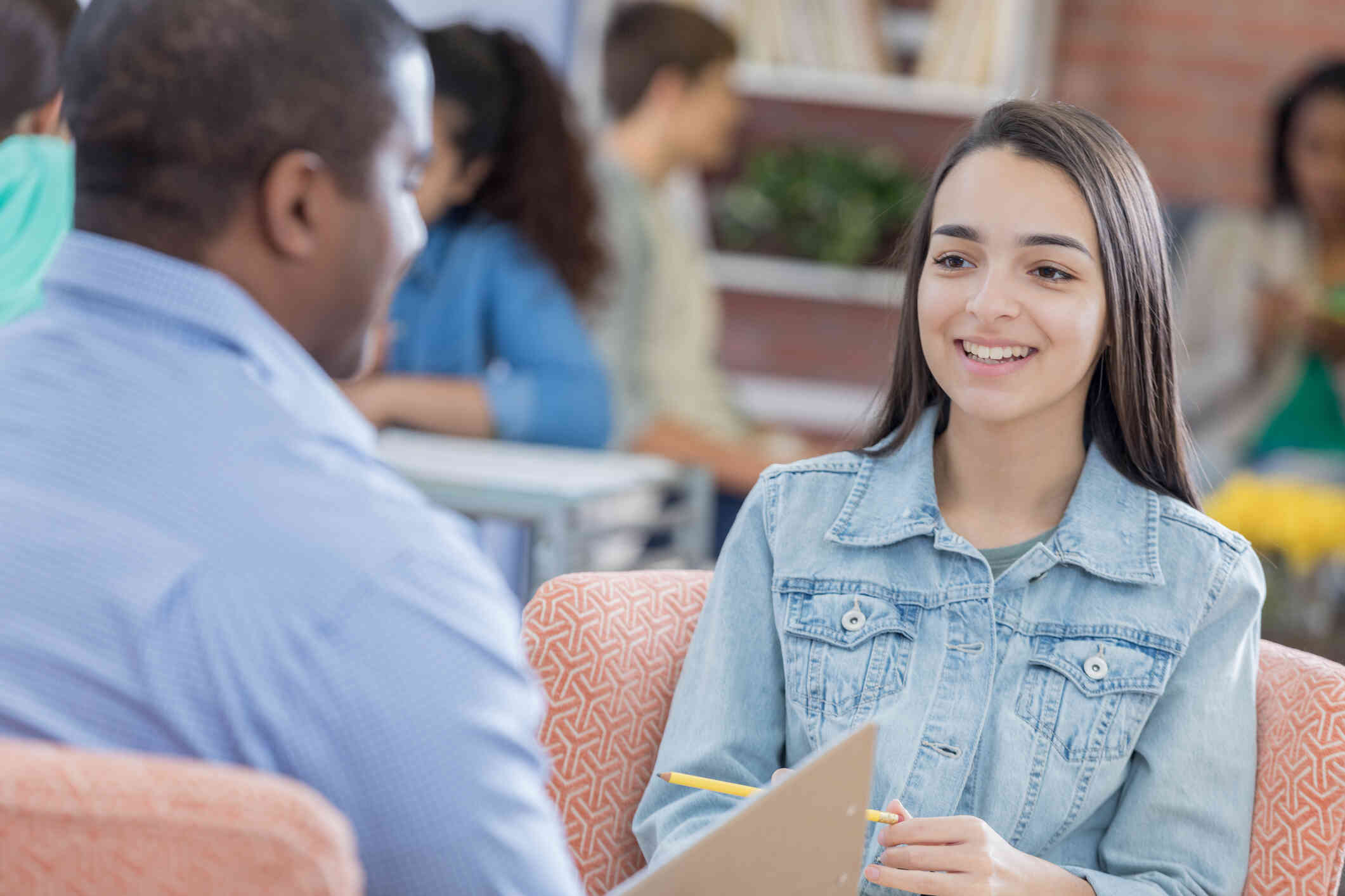 A young woman in a jean jacket smiles as she sits across from a man speaking to her in a crowded room.