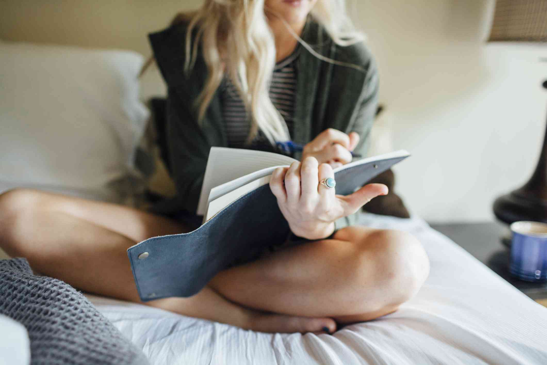 A girl with a dark green top is sitting cross legged on a bed write down notes and goals in her notebook