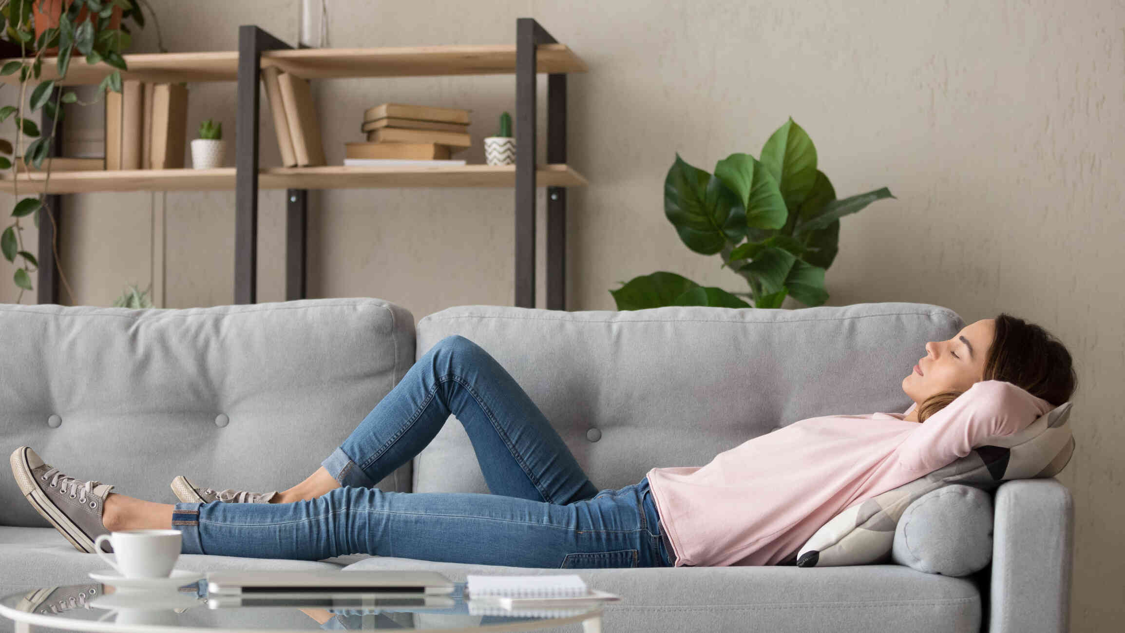 A woman in a pink shirt lays asleep  on her back on the couch with her hands behind her head.