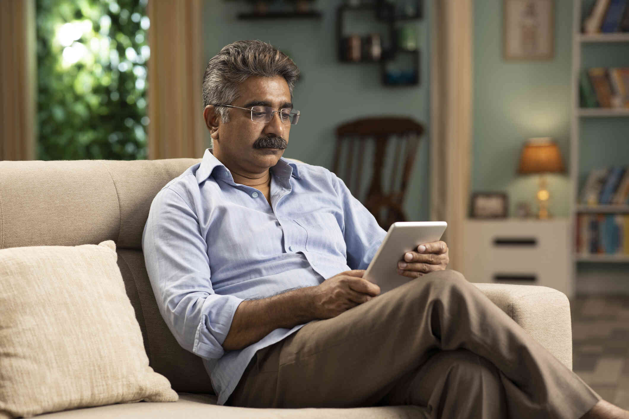 A man in a blue button down shirt sits on the couch and looks at the tablet in his hands.