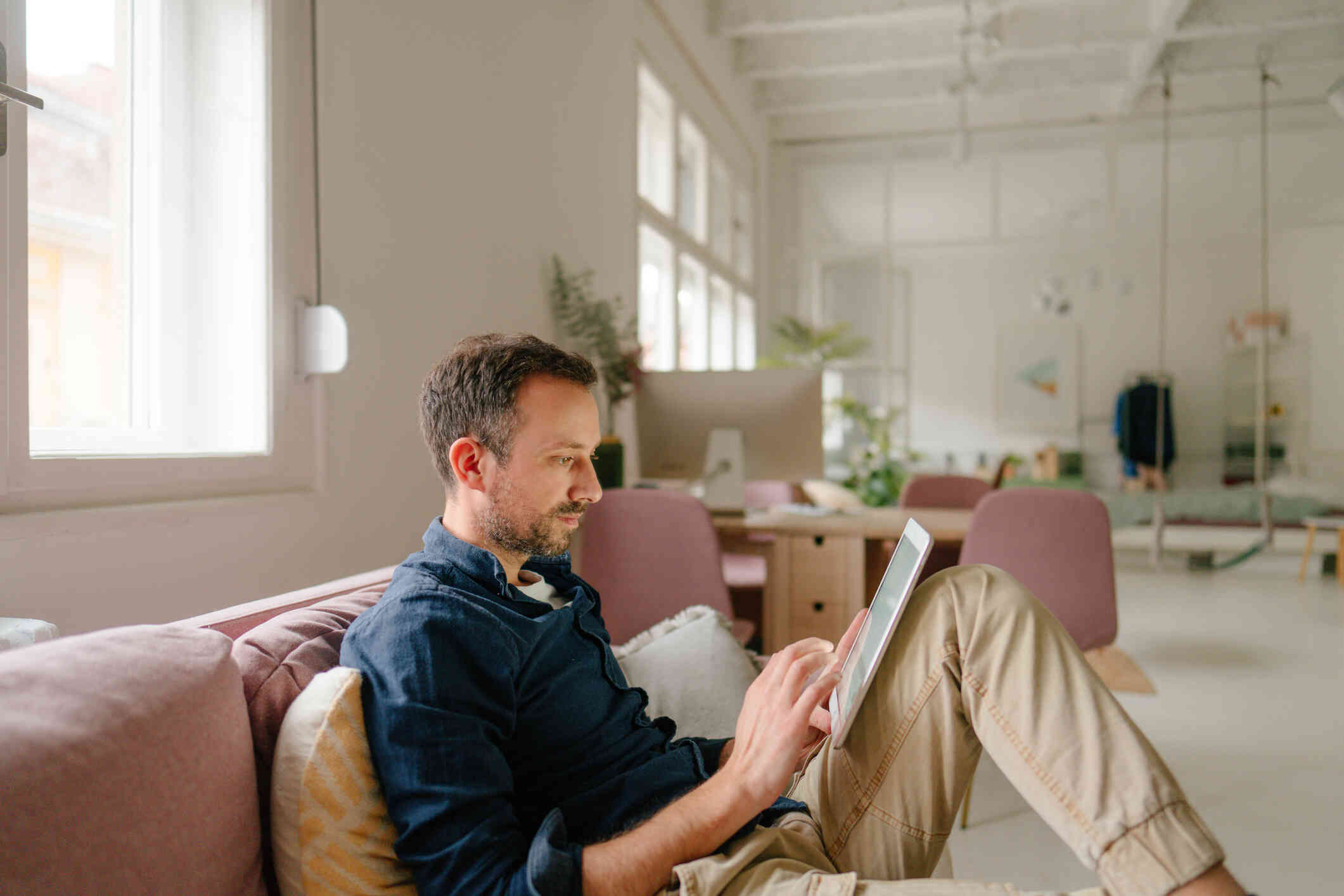 A man in a blue shirt sits casually on the couch and looks at the tablet resting against his leg as he taps on the screen.