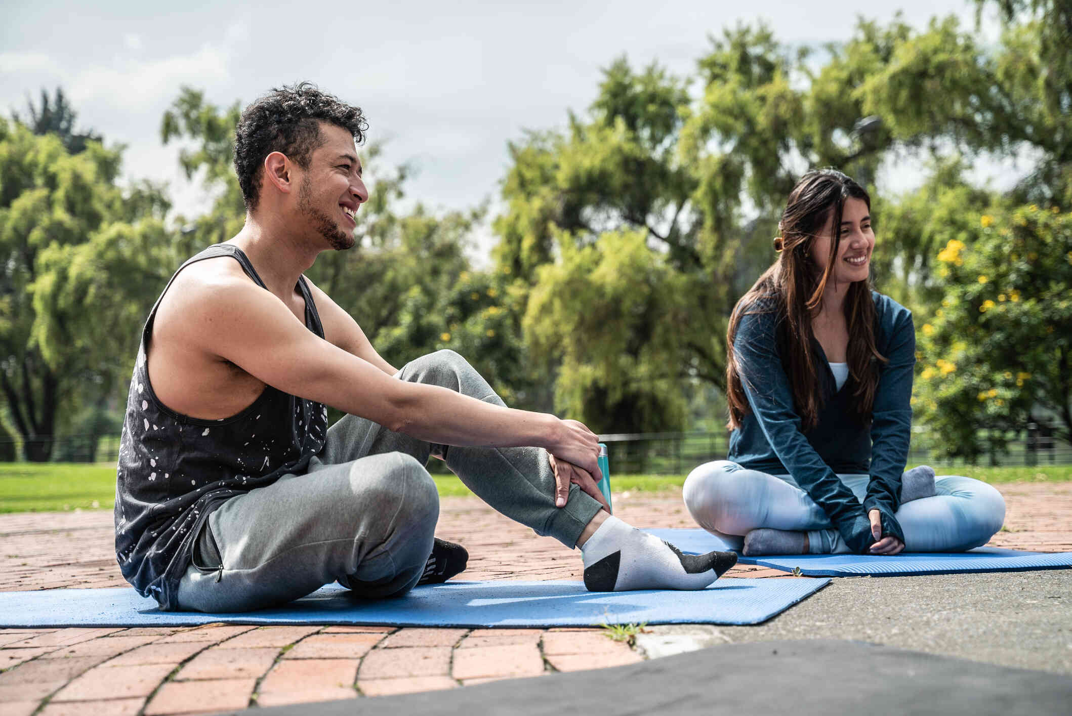 A man and his female friend sit on yoga mats next to each other outside on a sunny day.