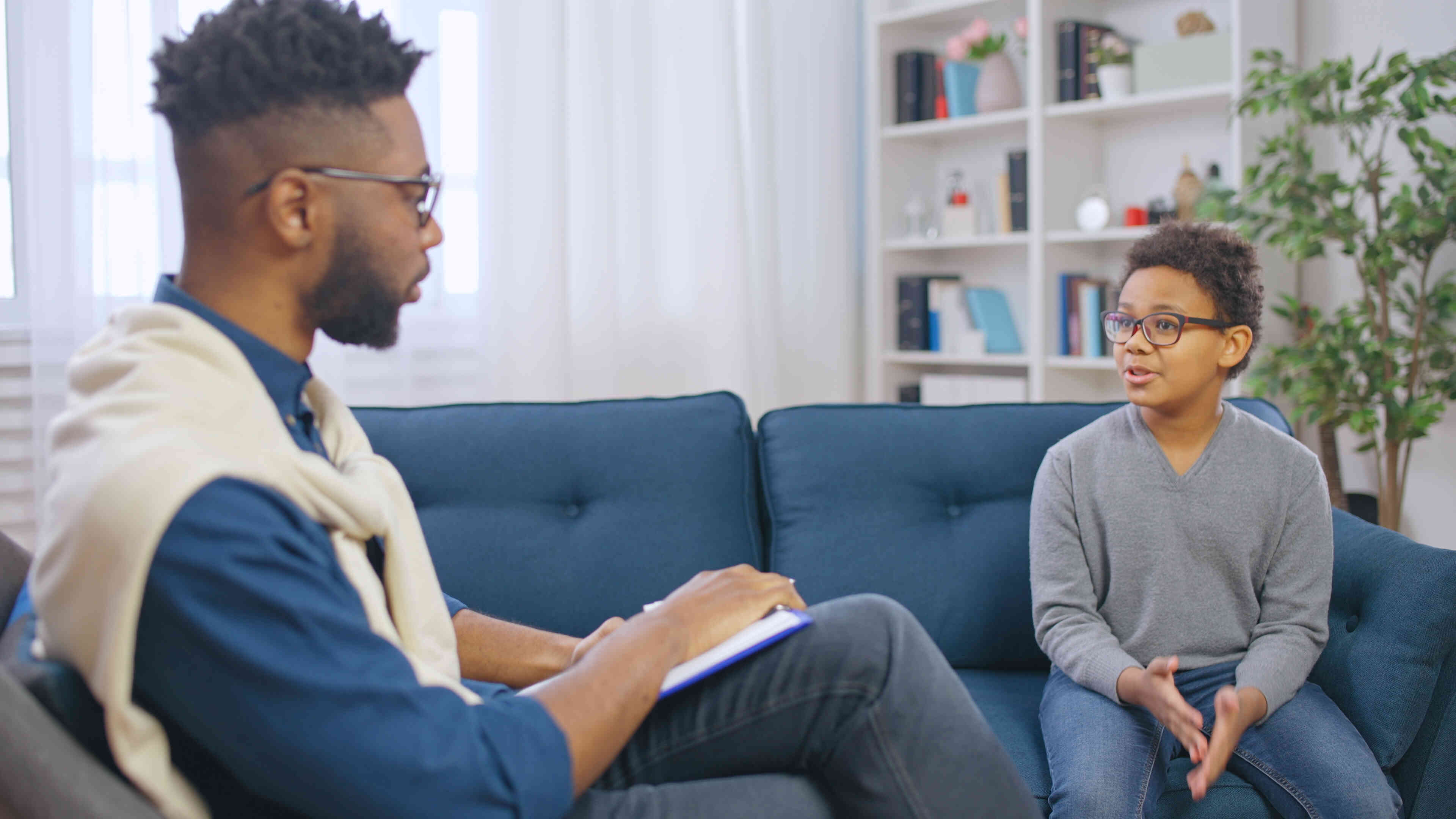 A young boy sits in on a blue couch and talks to the male therapist sitting in front of him.