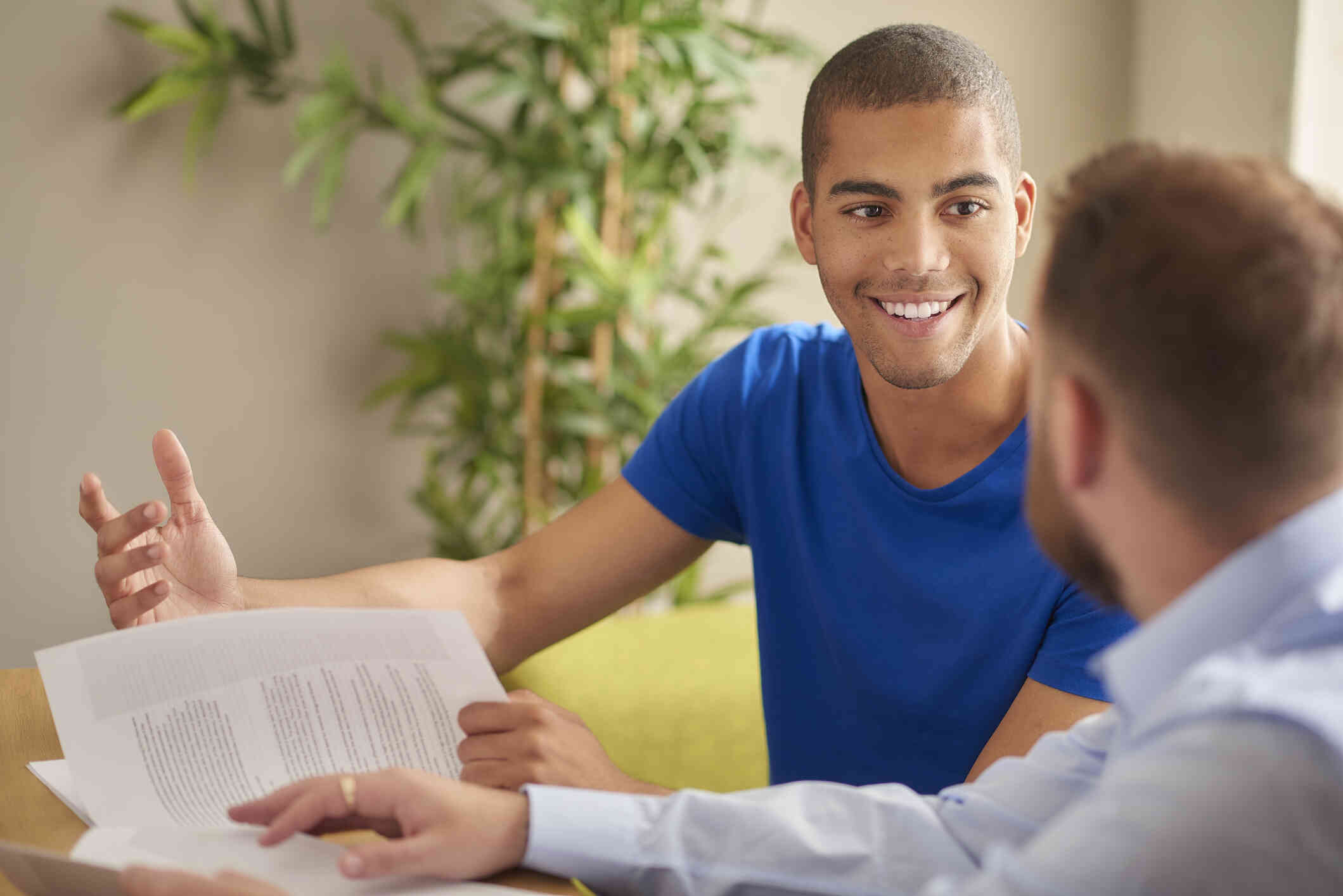 A man in a blue shirt sits at a table and holds some papers while smiling at the man sitting next to him.