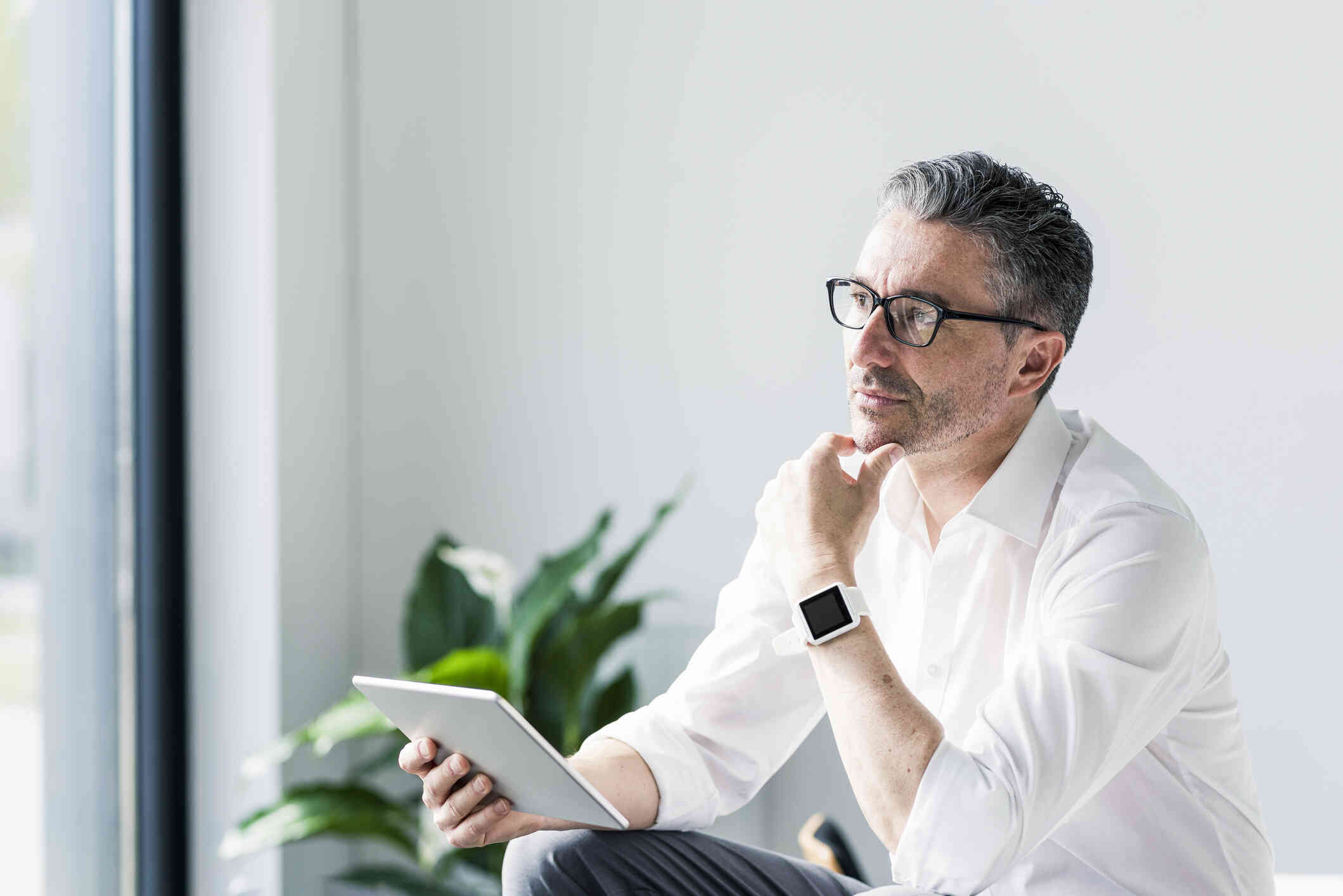 A middle aged man in a white button down shirt with  glasses sits at a table while holding his tablet and gazing off with a thoughtful expression.