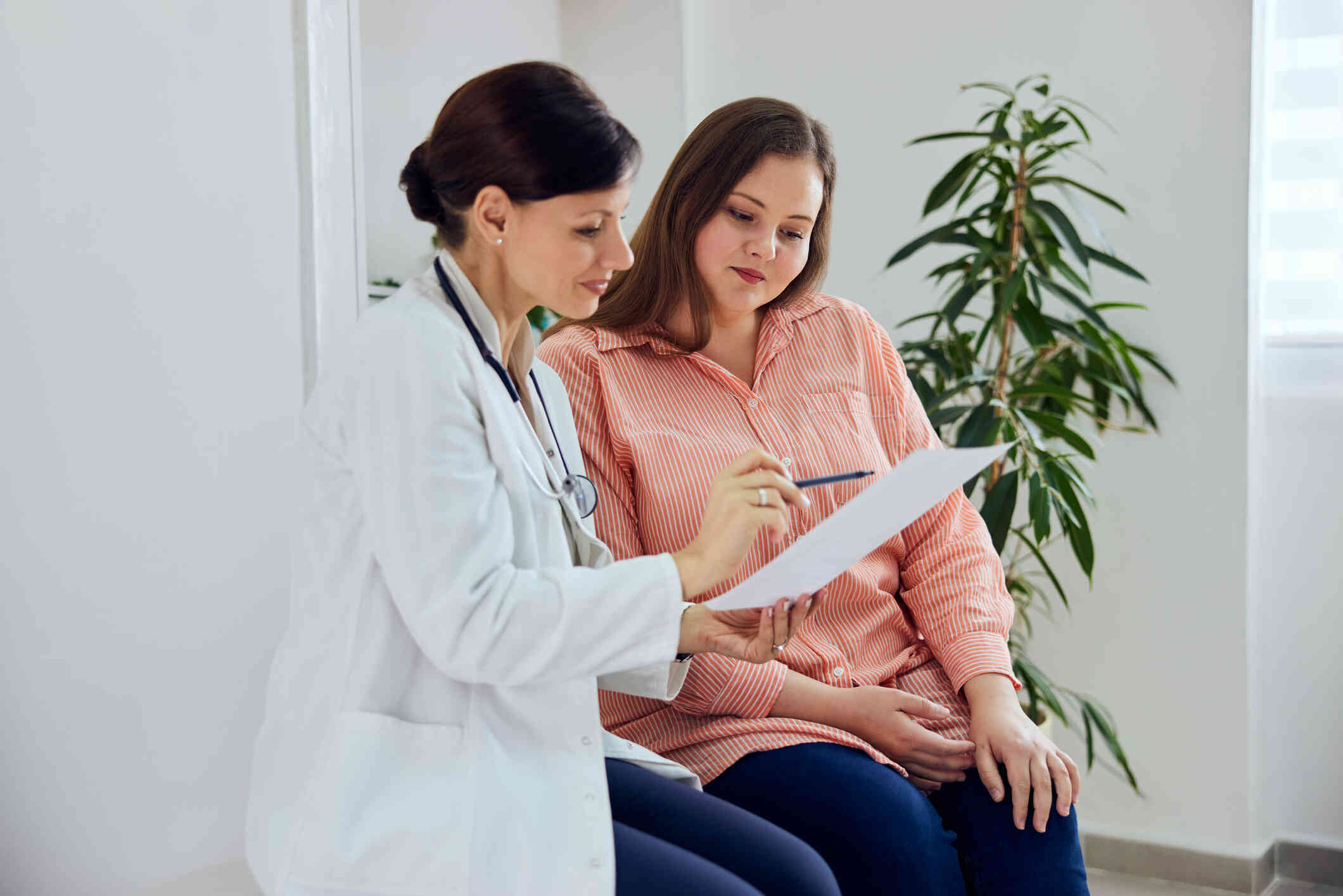 A woman in a pink top sits next to her female doctor while the doctor shows her some information on a piece of paper.