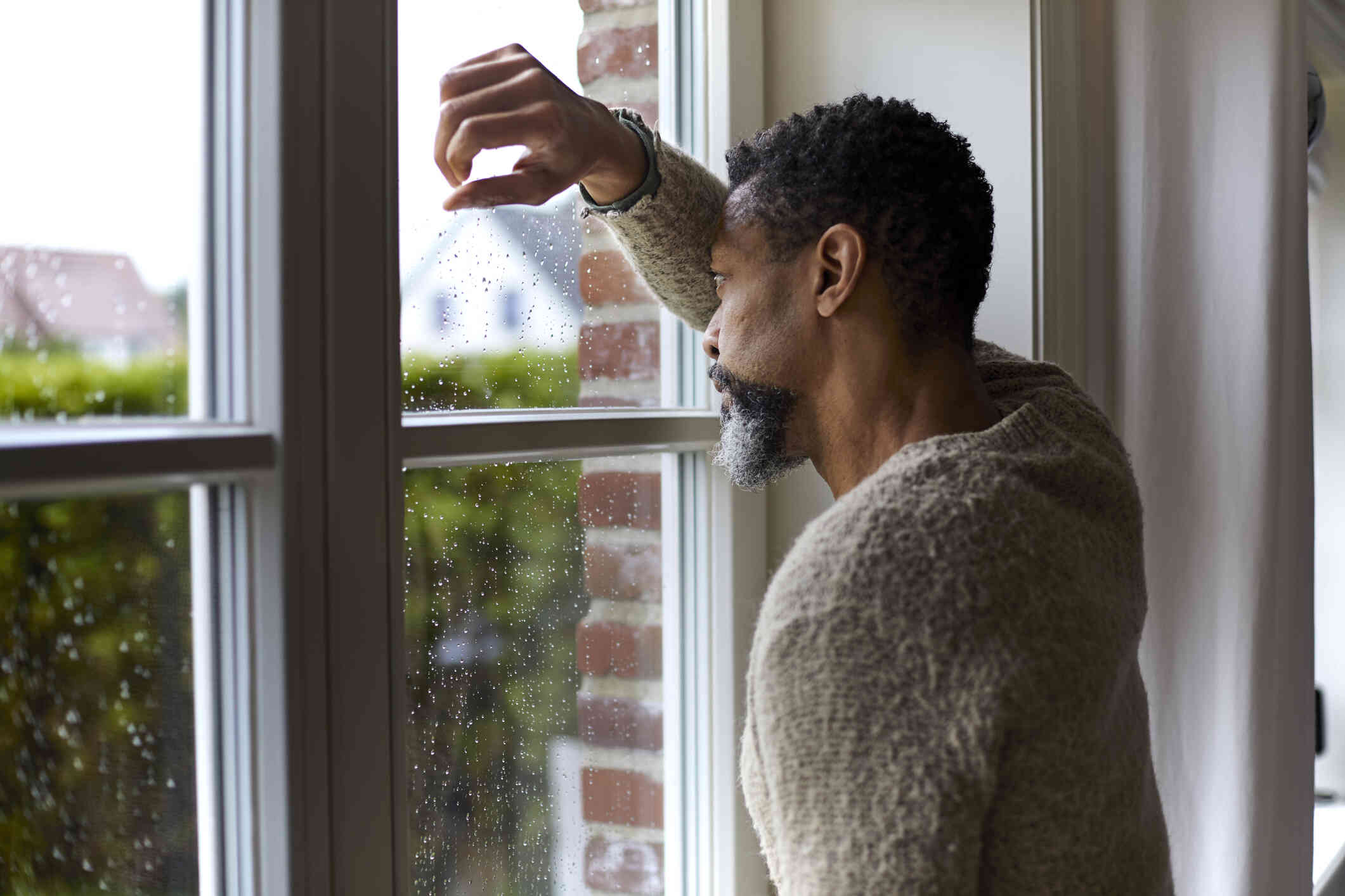 An elderly man in a sweater stands at the window and rests his head against his arm while gazing out with a sad expression.