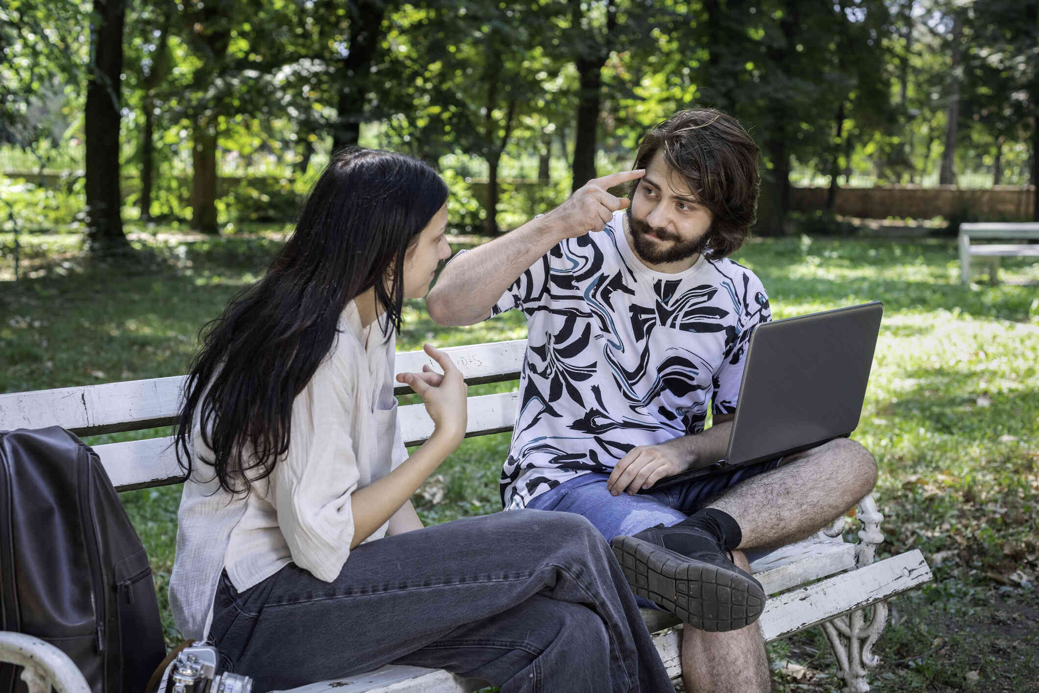 A woman and her male friend sit on a park bench on a sunny day and talk using sign language.