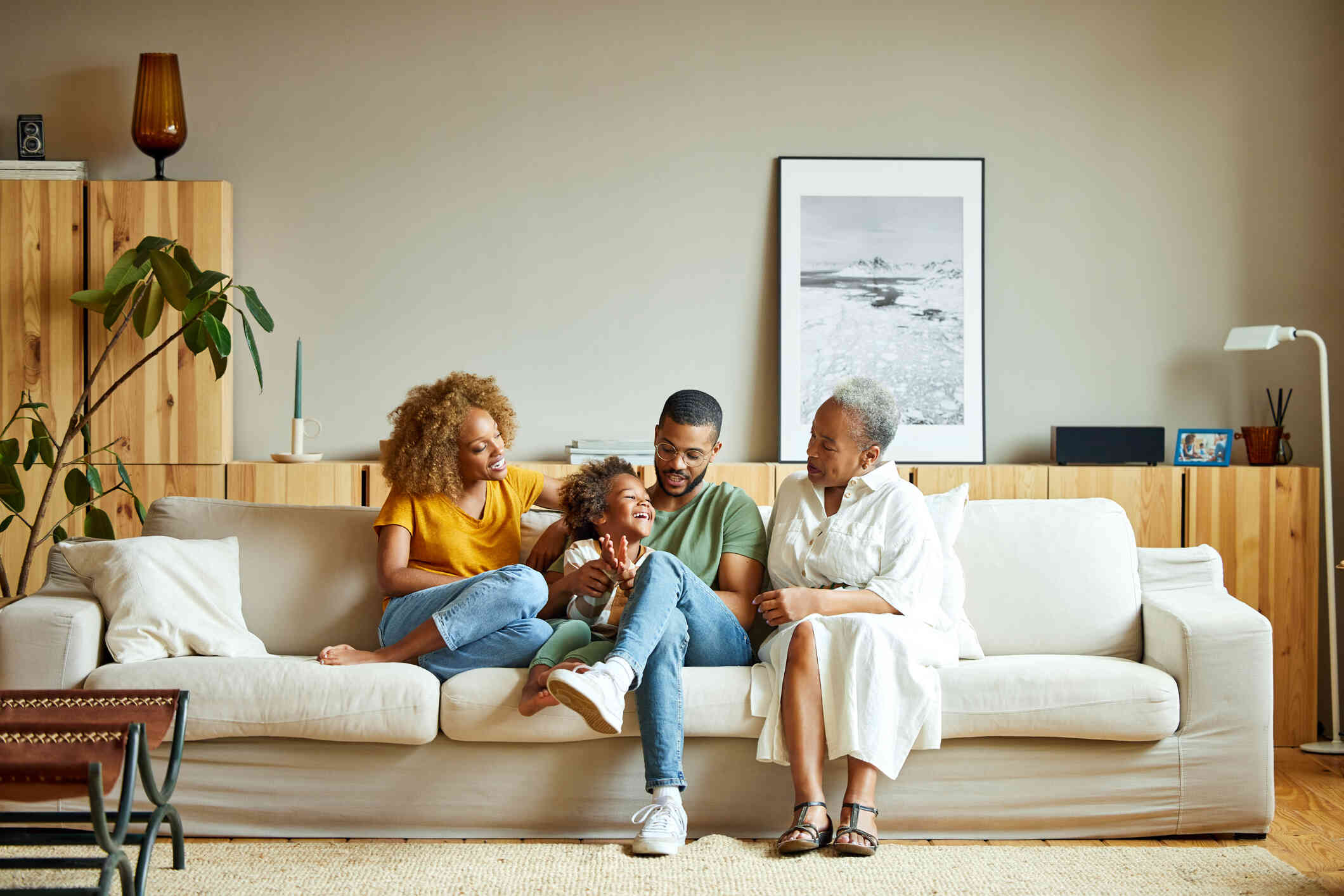 A mother, father, grandmother, and young daughter happily sit on a couch together while laughing.