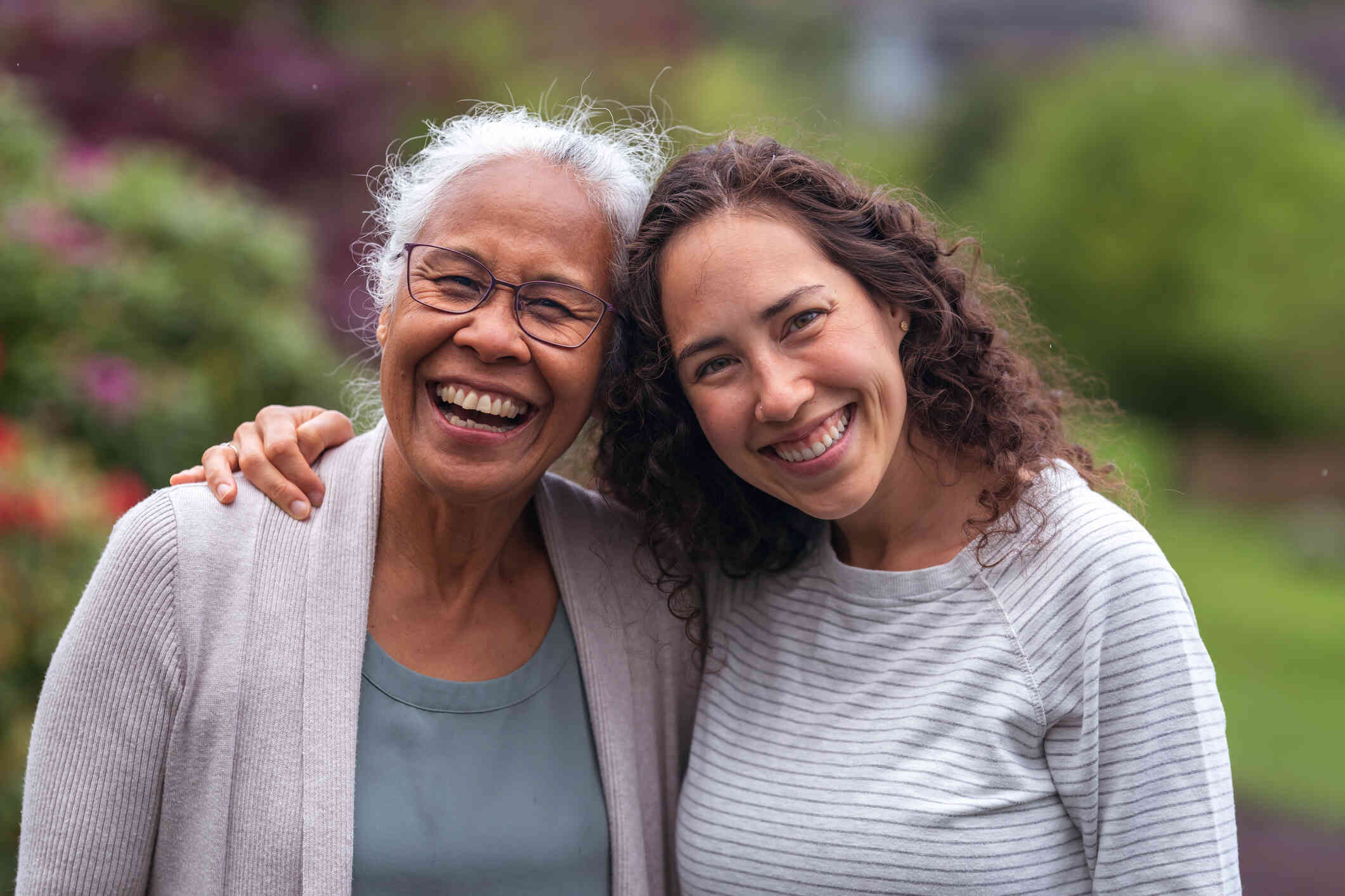 A woman wraps her arm around an older womans shoulder as they both smile and look at the camera.