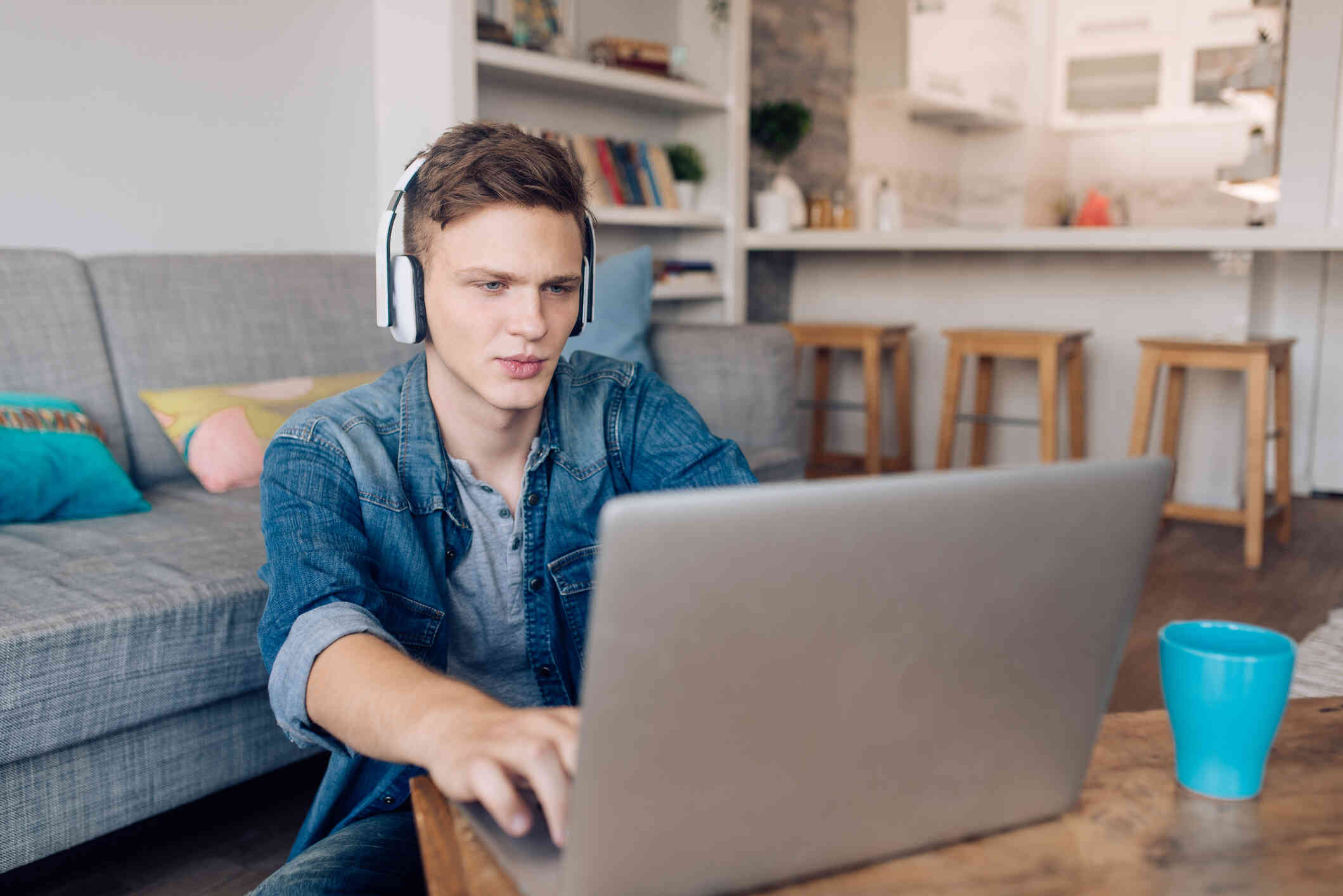 A teen boy wearing a blue jean shirt and headphones sits at a table with his laptop open infront of him and looks at the screen with a focused expression.
