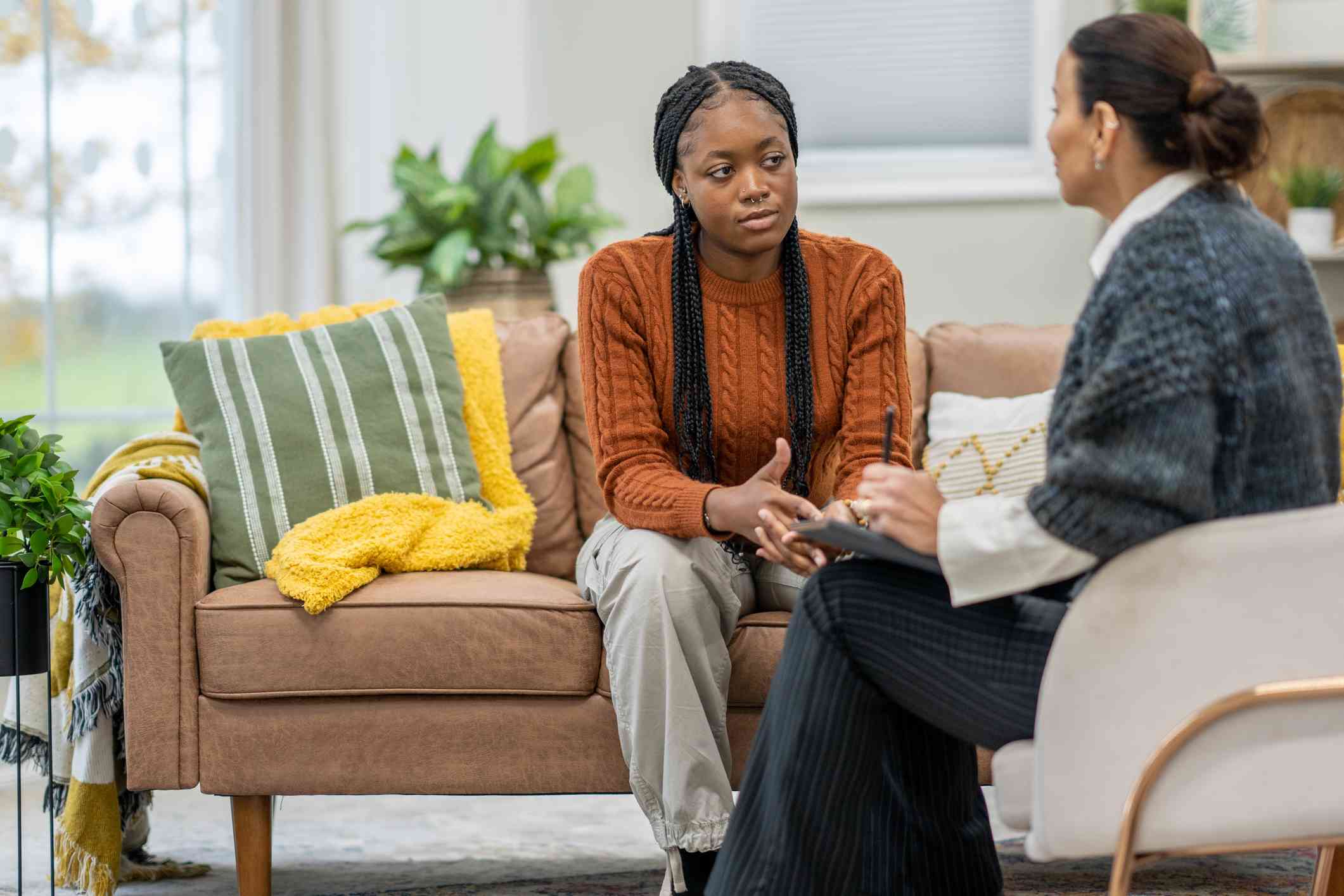 A woman in an orange sweater sits hunched over on a couch and talks to the female therapist that sits infront of her with a worried expression.