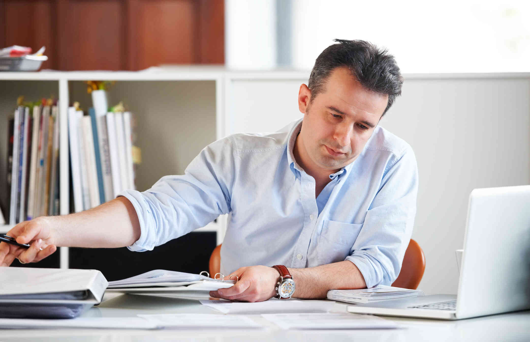 A man sits at a table at work with lots of papers spread out infront of him along with an open laptop as he sorts through papers with a serious expression.