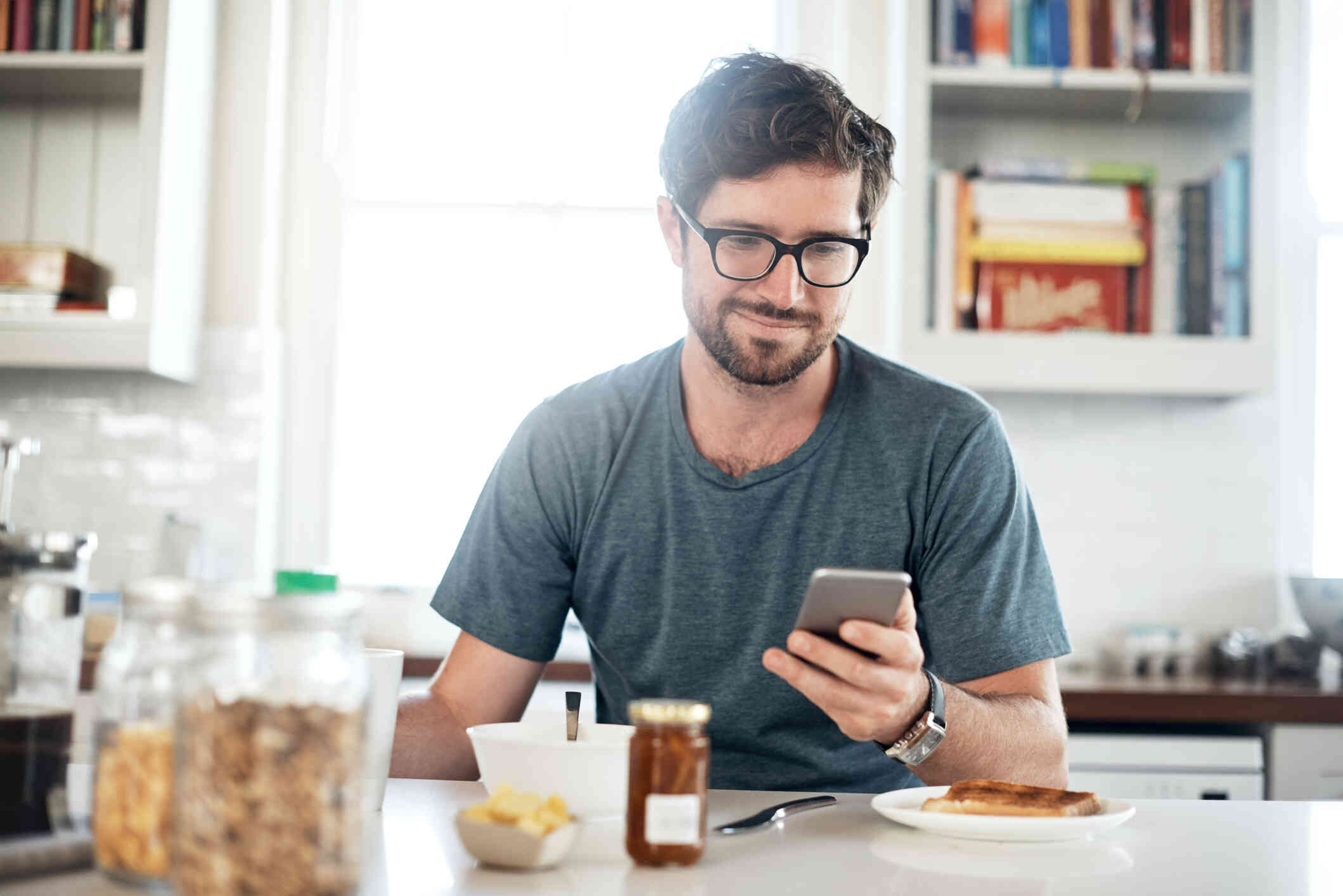 A man with glasses sits at the kitchen table with his breakfast and looks down at the cellphone in his hand with a soft smile.