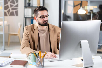 A man with a dark beard and glasses sits at a desk with a focused expression and looks at a desktop computer screen.