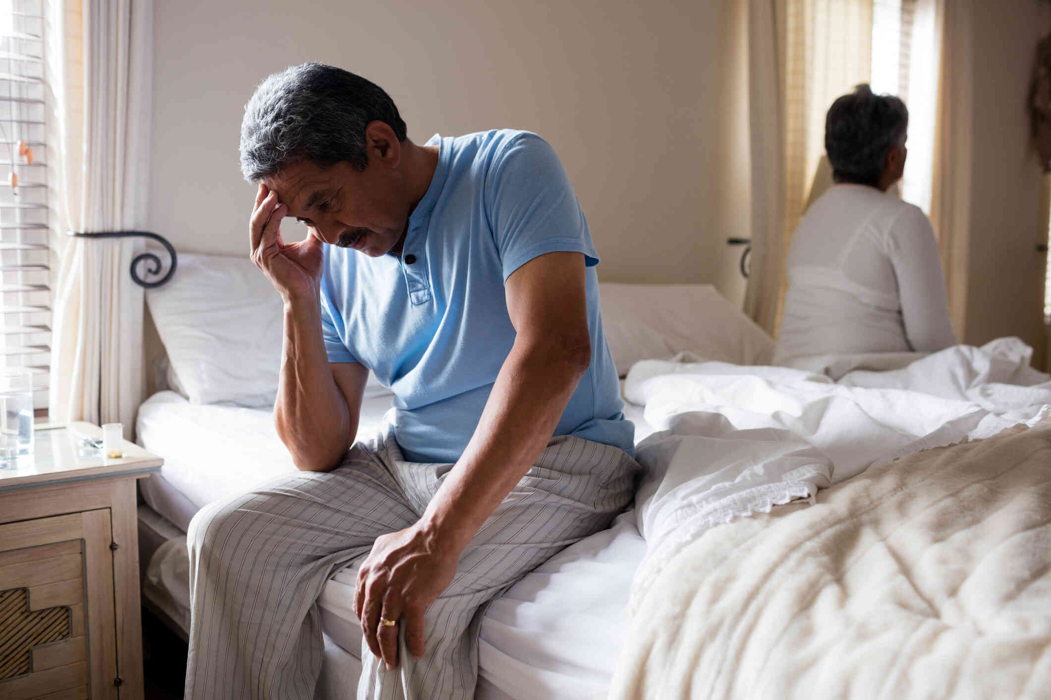 A man sits on the edge of a couch with his hands to his face looking distressed while his wife sits in the background gazing off